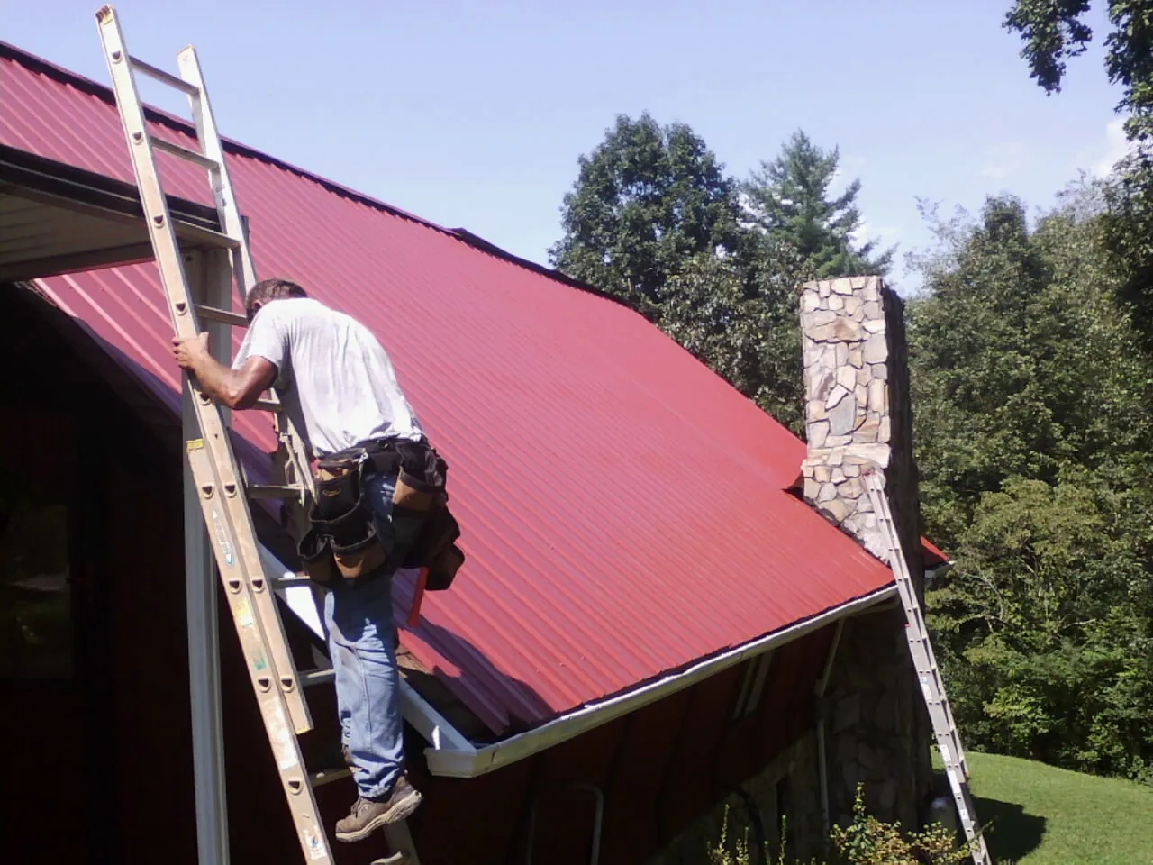 Roofer on a ladder beside a red metal roof and stone chimney — Soffit & Fascia Repair by Roofing Brothers in Yadkinville